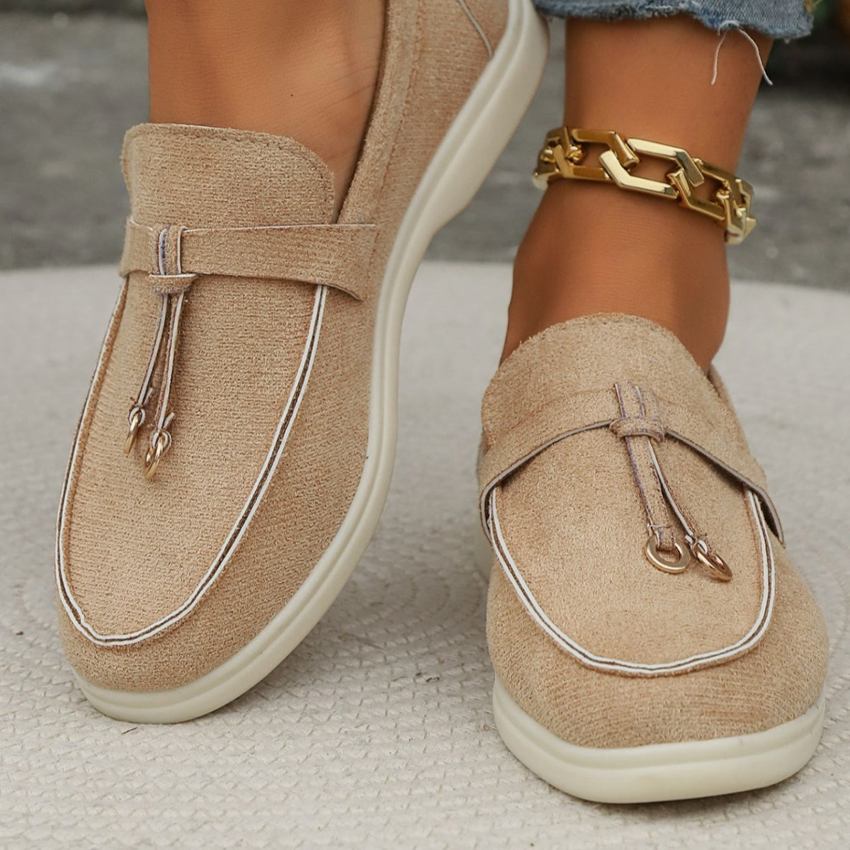 Khaki flat slip-ons with decorative strap and white piping detail.