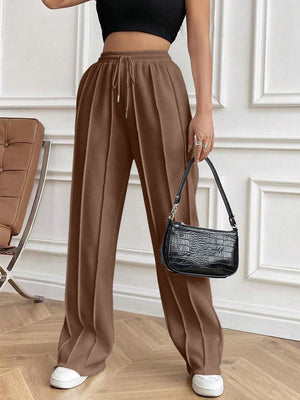 Brown pleated palazzo trousers with drawstring waist and wide leg, styled with a black shoulder bag.