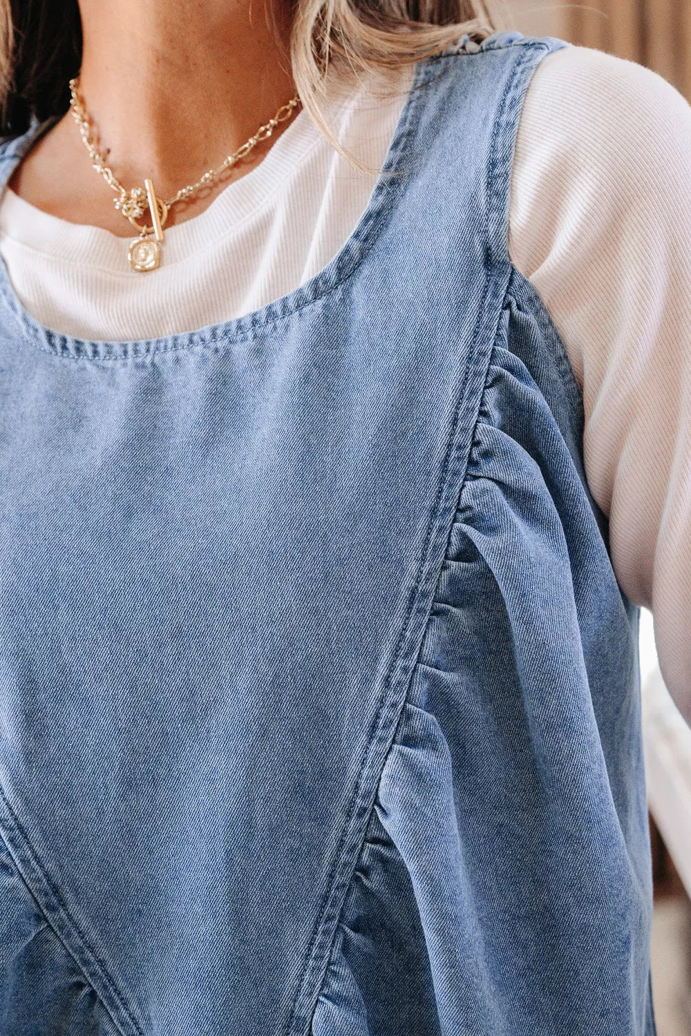 V-shaped pleated denim sleeveless top with square neckline, styled with a white long-sleeve shirt.