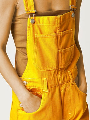 Gold denim short overalls with crisscross straps and front pockets.
