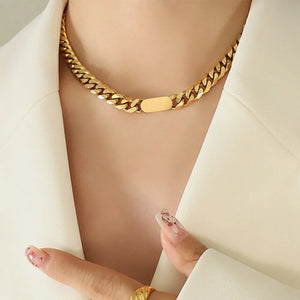 Gold Cuban link necklace with a flat oval accent, worn with a white blazer.