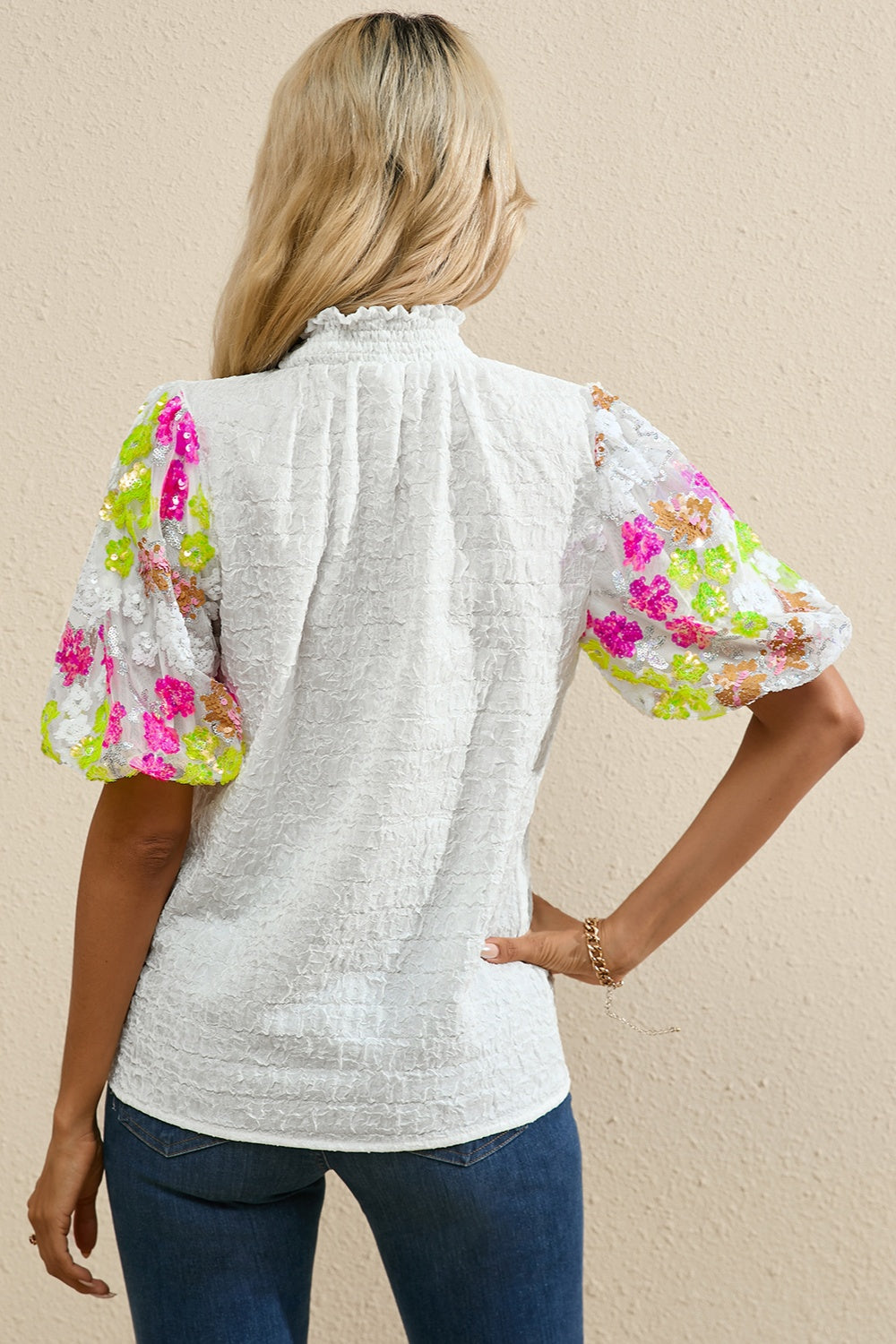 White textured blouse with puff sleeves featuring colorful sequin floral detail and ruffled neckline.