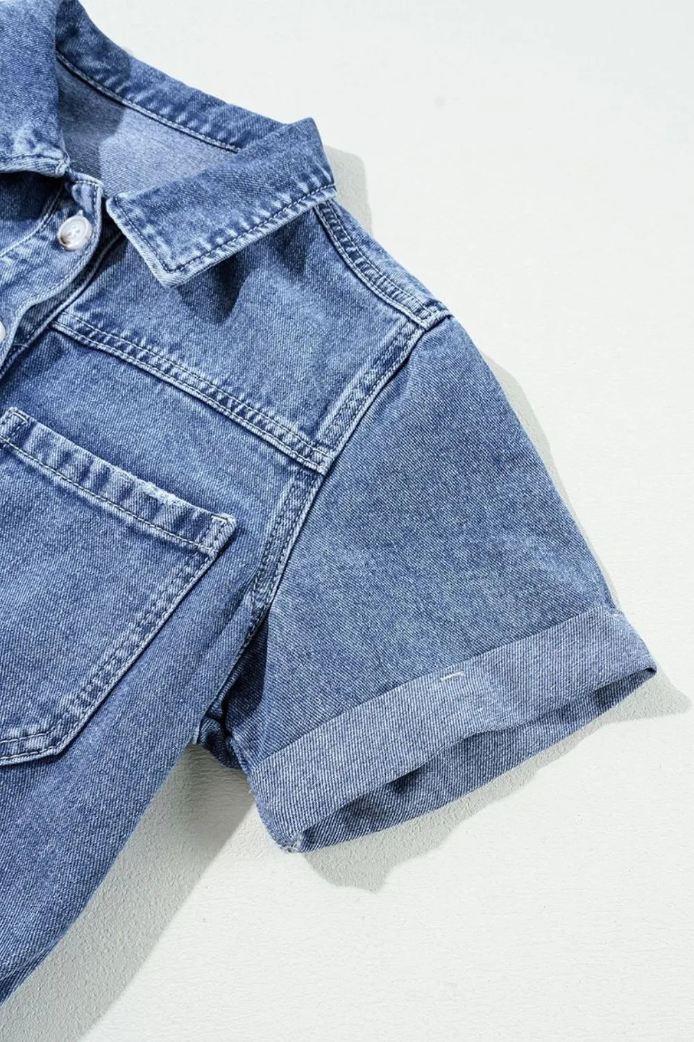 Medium wash denim romper with short sleeves, button-down front, chest pockets, and cuffed hem.