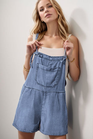 Medium wash denim romper with adjustable straps and a front pocket.
