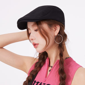 Black cotton newsboy cap with a flat brim, worn by a model with braided hair.