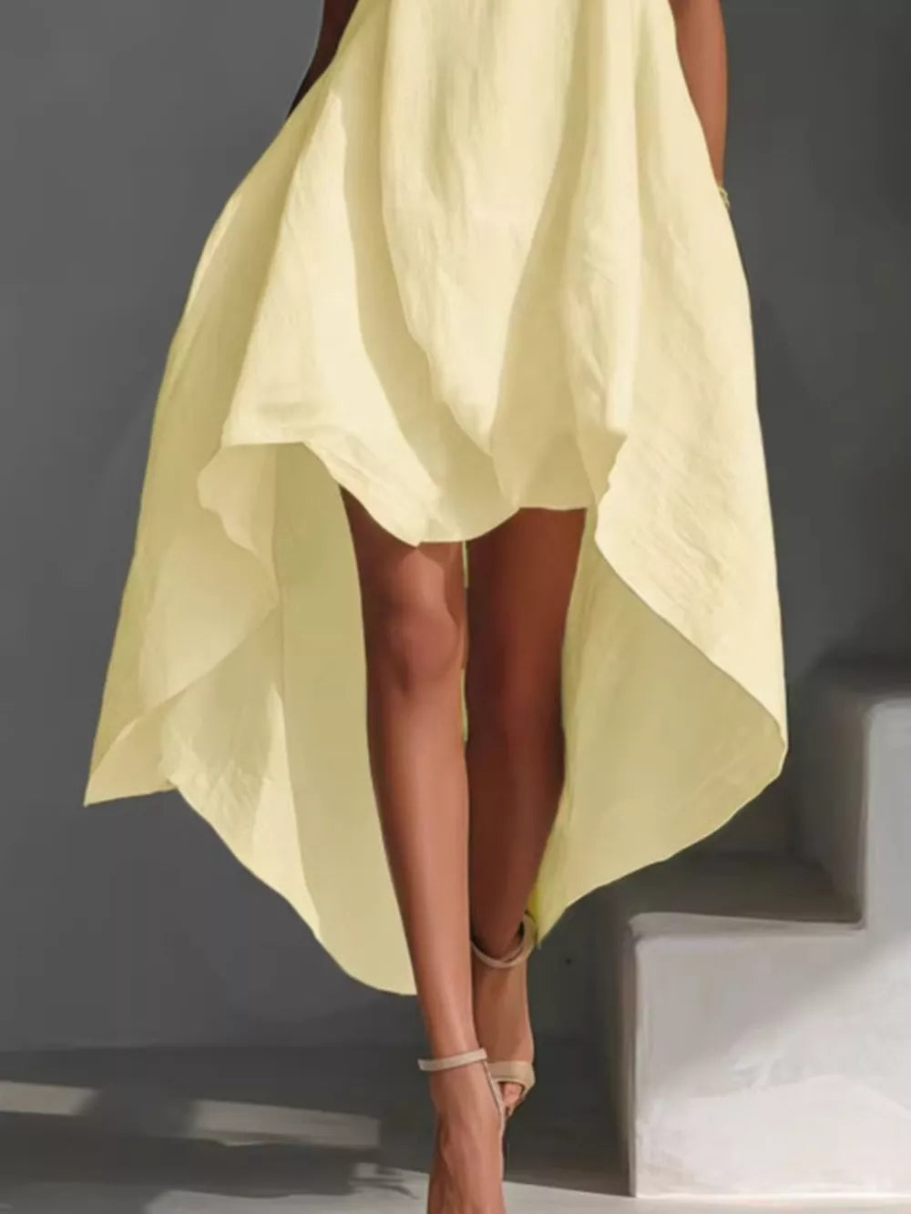 Pastel yellow asymmetrical sleeveless flowing dress with high-low hem and strappy heels.