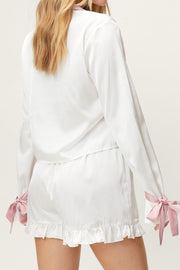 White silk pajama set with pink bow details on sleeves and ruffled shorts.