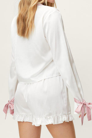 White silk pajama set with pink bow details on sleeves and ruffled shorts.