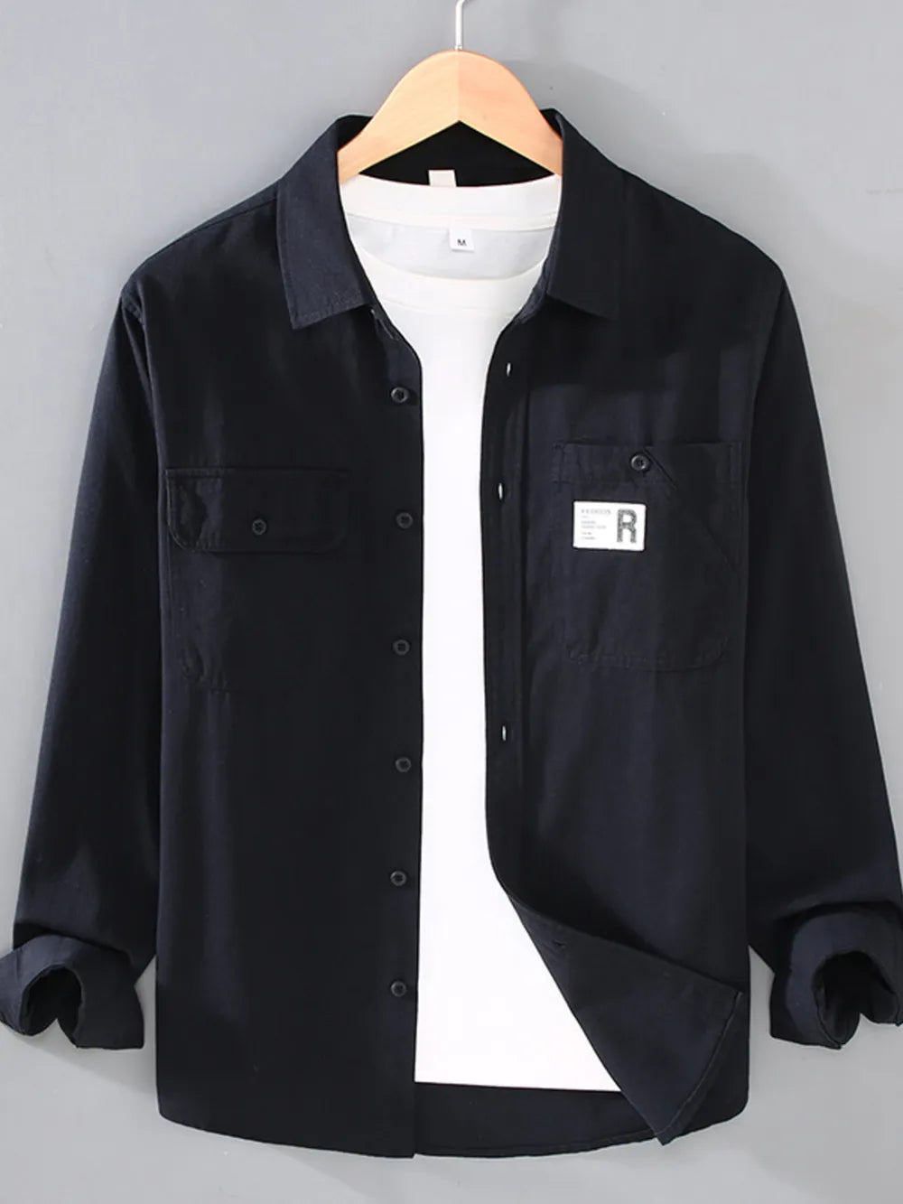 Black cotton long-sleeve overshirt with two chest pockets and button closure, worn over a white t-shirt.