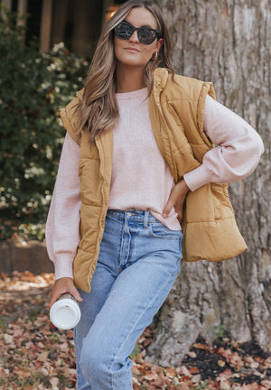 Tan padded hooded vest with pockets, sleeveless, worn over a pink sweater and jeans.