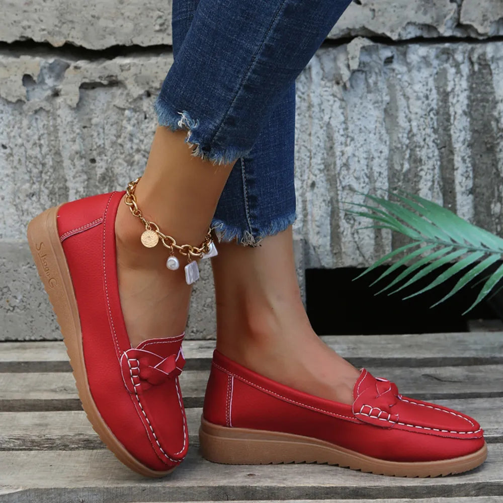 Red leather loafers with braided detail and white stitching, worn with jeans.