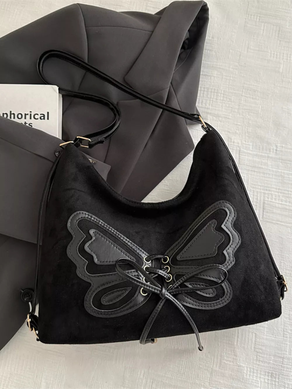 Black suede half-moon shoulder bag with butterfly embellishment and lace-up detail.