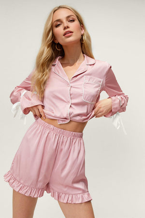 Blush pink silk pajama set with white trim, featuring a button-up top with tie sleeves and ruffled shorts.