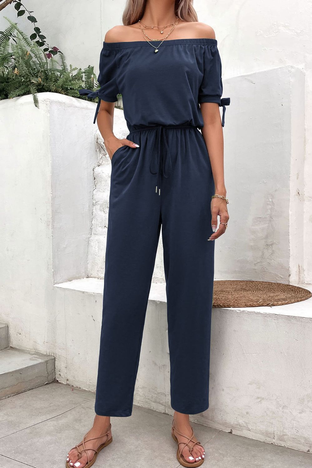 Off-the-shoulder navy jumpsuit with tie sleeves, drawstring waist, and pockets.