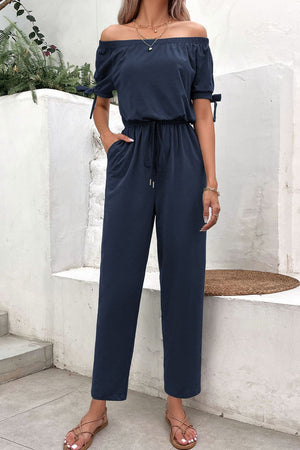 Off-the-shoulder navy jumpsuit with tie sleeves, drawstring waist, and pockets.