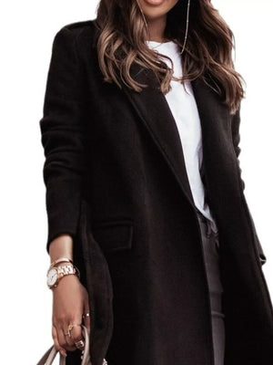 Black longline trench coat with flap pockets and a belt, worn over a white top.