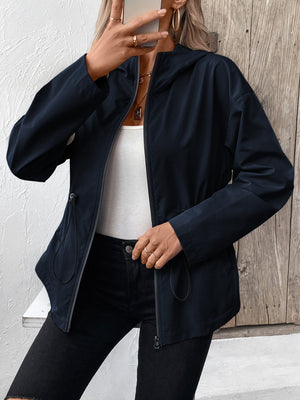 Navy utility jacket with cinched waistline, hood, and zip front, styled with a white tank and black jeans.
