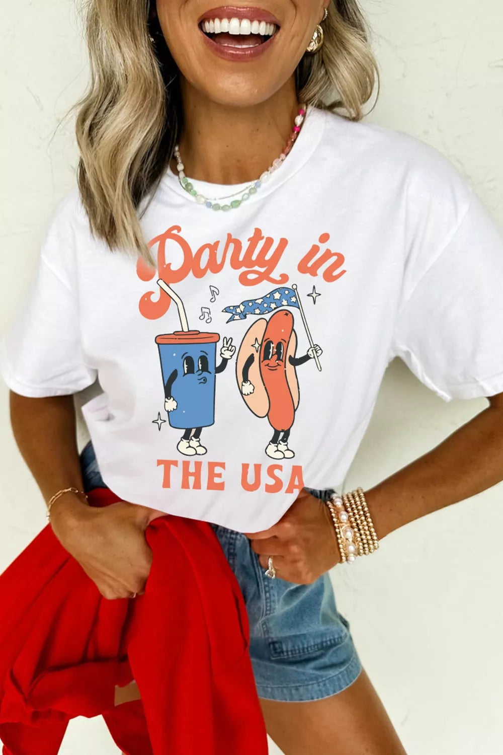 White graphic t-shirt with patriotic cartoon cup and hot dog, "Party in the USA" text.