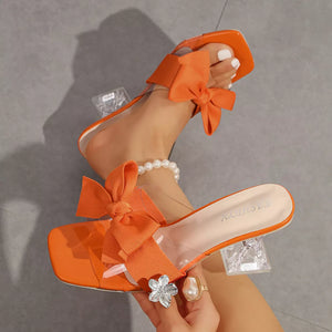 Orange open-toe sandals with clear straps and large bows, featuring a block heel.