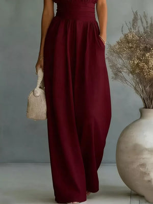 Burgundy sleeveless wide-leg jumpsuit with cinched waist and pockets, styled with a woven bag.