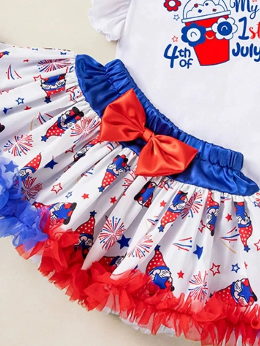 Baby girl's festive outfit with gnome and firework print skirt, red bow, and ruffled hem.