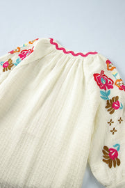 Beige textured blouse with colorful floral embroidery on sleeves and pink zigzag neckline.