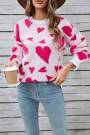 Pink intarsia heart knit jumper with crew neck and long sleeves, styled with jeans.