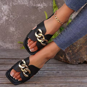 Black slide sandals with gold chain detail and square toe, worn with jeans.