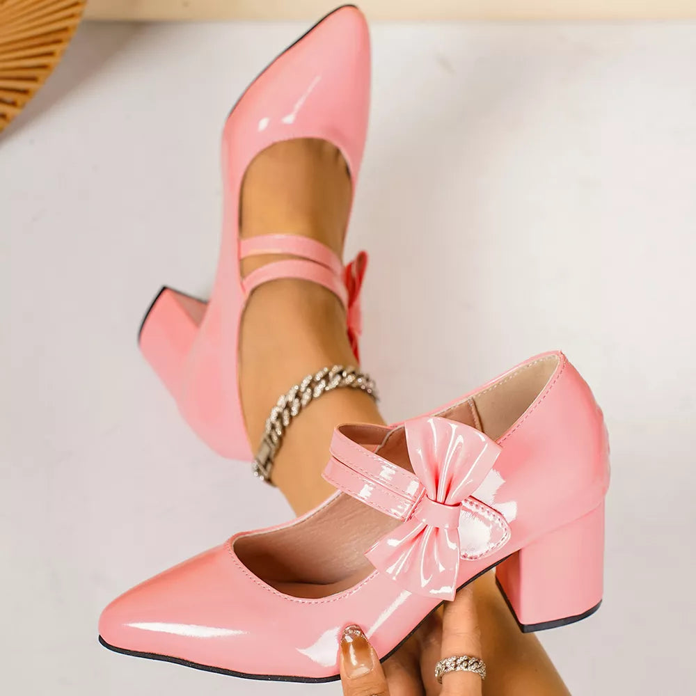 Pink patent pointed toe block heel pumps with ankle straps and bow detail.