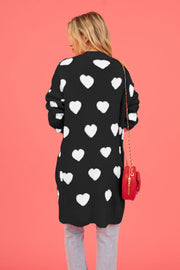 Black longline knit cardigan with allover white heart motif, crew neck, and long sleeves.