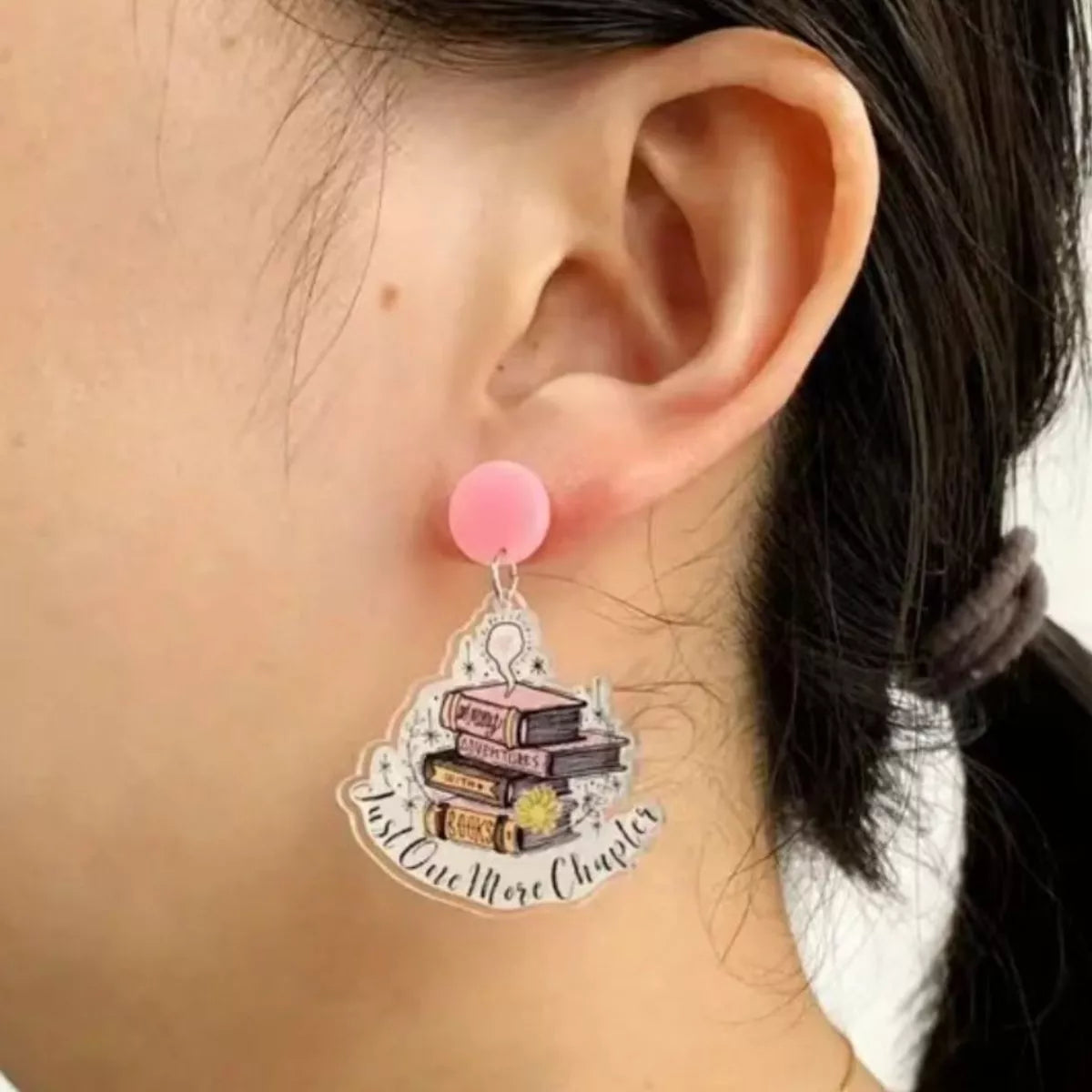 Blush pink acrylic drop earrings with stacked books and "Just One More Chapter" text.