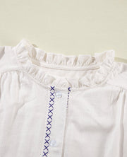 White linen blouse with ruffled neckline and navy cross-stitch embroidery detail.
