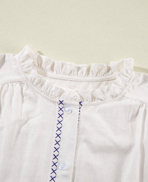 White linen blouse with ruffled neckline and navy cross-stitch embroidery detail.