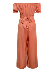 Coral off-the-shoulder jumpsuit with ruched bodice, short puff sleeves, and wide-leg pants cinched with a self-tie sash.