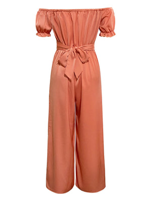 Coral off-the-shoulder jumpsuit with ruched bodice, short puff sleeves, and wide-leg pants cinched with a self-tie sash.