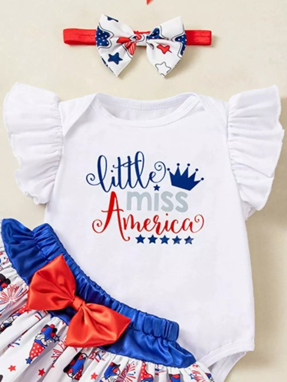White baby outfit with ruffled sleeves and "little miss America" graphic. Includes patriotic headband and skirt.