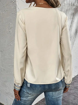 Beige V-neck blouse with embroidered trim on cuffs, long sleeves, paired with distressed jeans.