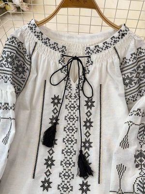 White embroidered tie neck blouse with black tassels and long sleeves.