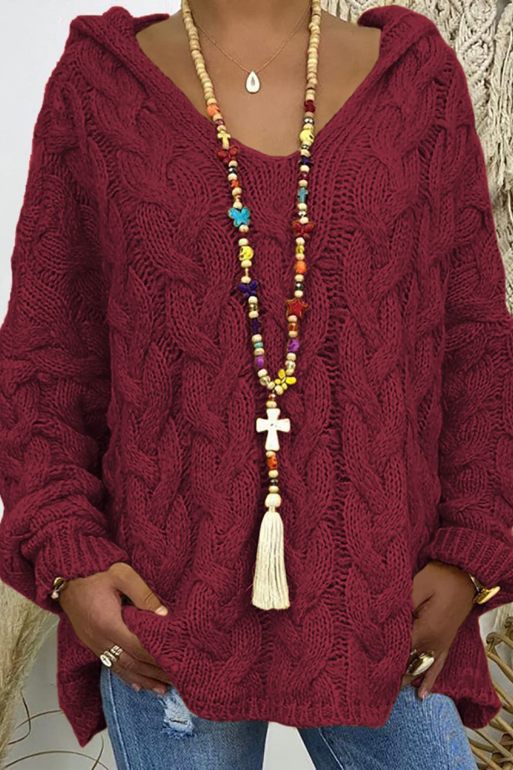 Burgundy cable knit hooded cocoon sweater with V-neck and tassel necklace.
