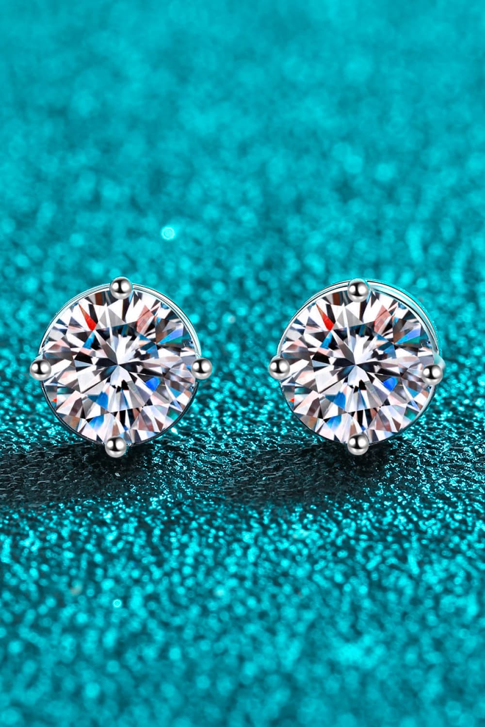 Moissanite solitaire stud earrings with rhodium finish on a teal textured background.