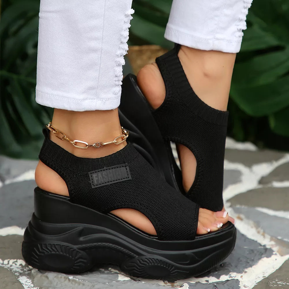 Black open-toe wedge sandals with chunky sole and ankle chain detail.