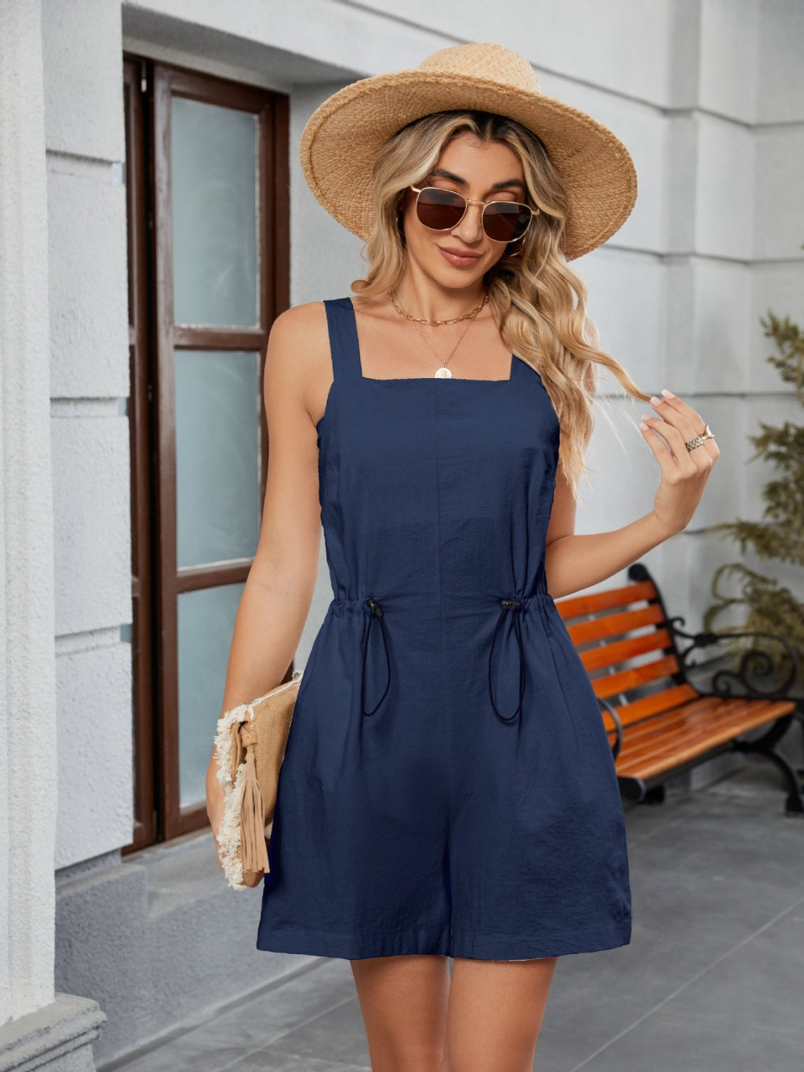 Navy square-neckline romper with drawstring waist and pockets, styled with a straw hat and clutch.
