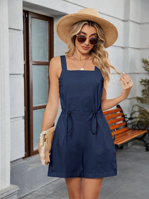 Navy square-neckline romper with drawstring waist and pockets, styled with a straw hat and clutch.