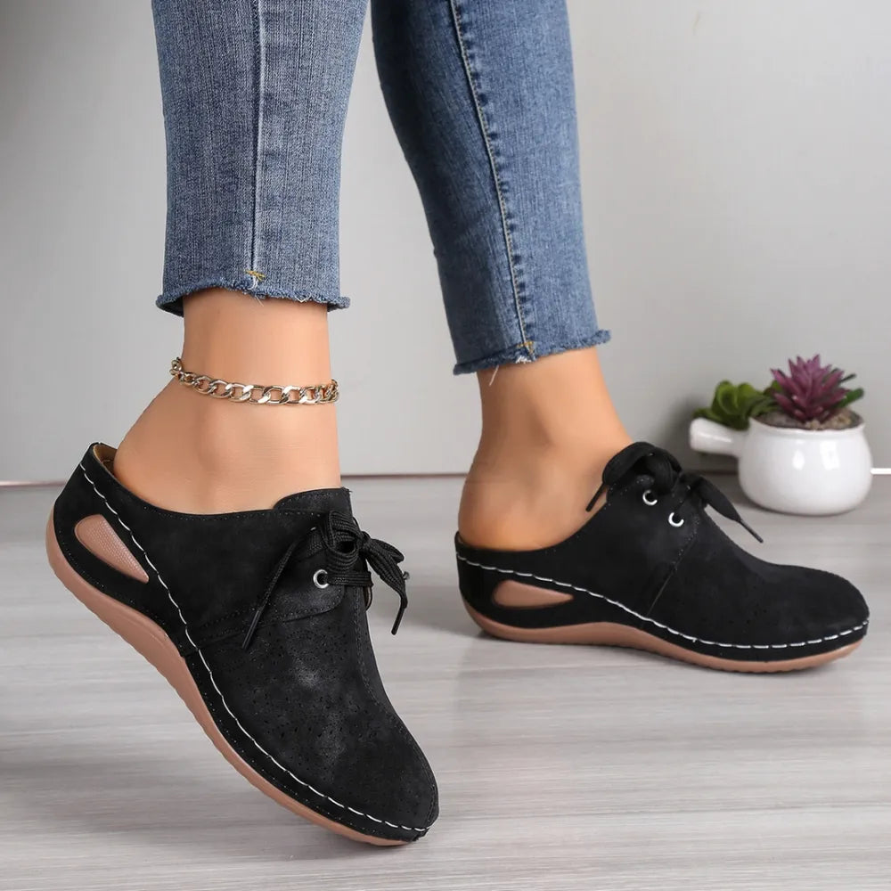 Black perforated lace-up mule flats with cutout sides and white stitching detail.