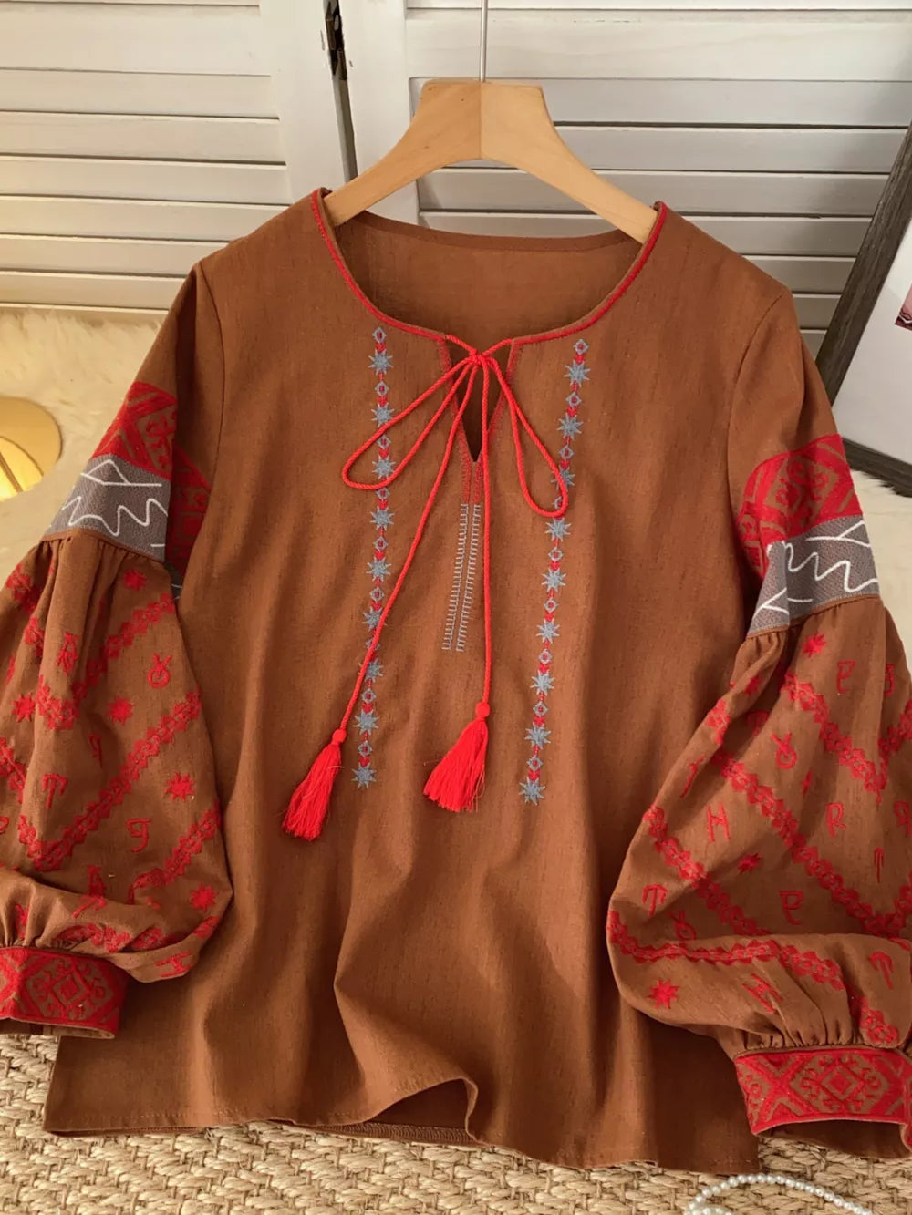 Brown folk-inspired tie neck shirt with red embroidered sleeves and tassels.