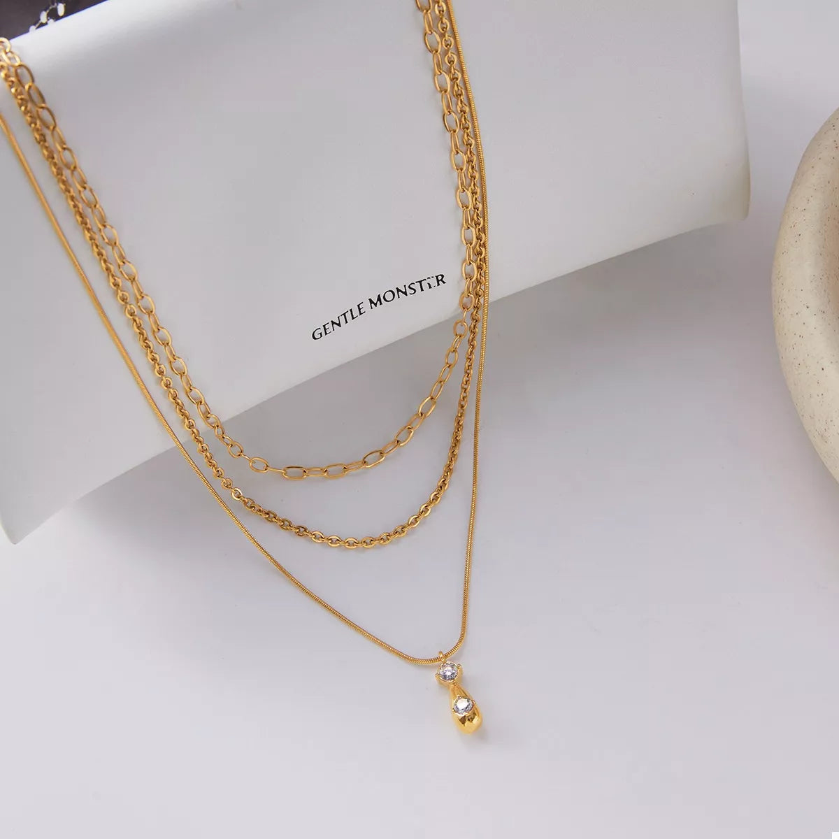 Gold three-layered pendant necklace with mixed chains and a small clear stone accent.