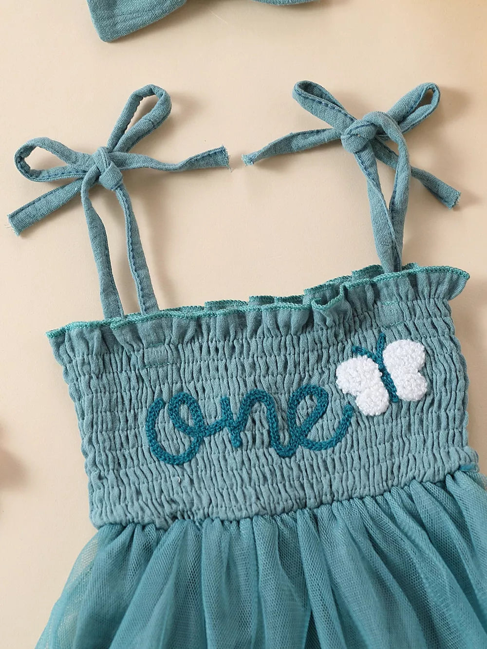 Sea blue smocked baby bodysuit with embroidered "one" and butterfly detail, tie straps.