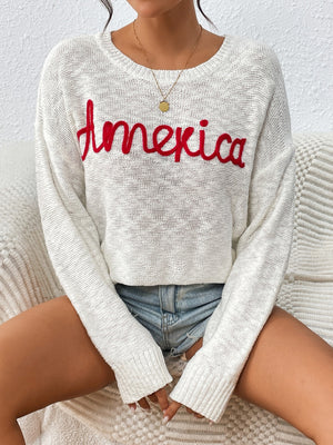 Ivory knit sweater with red embroidered "America" text, crew neck, long sleeves.