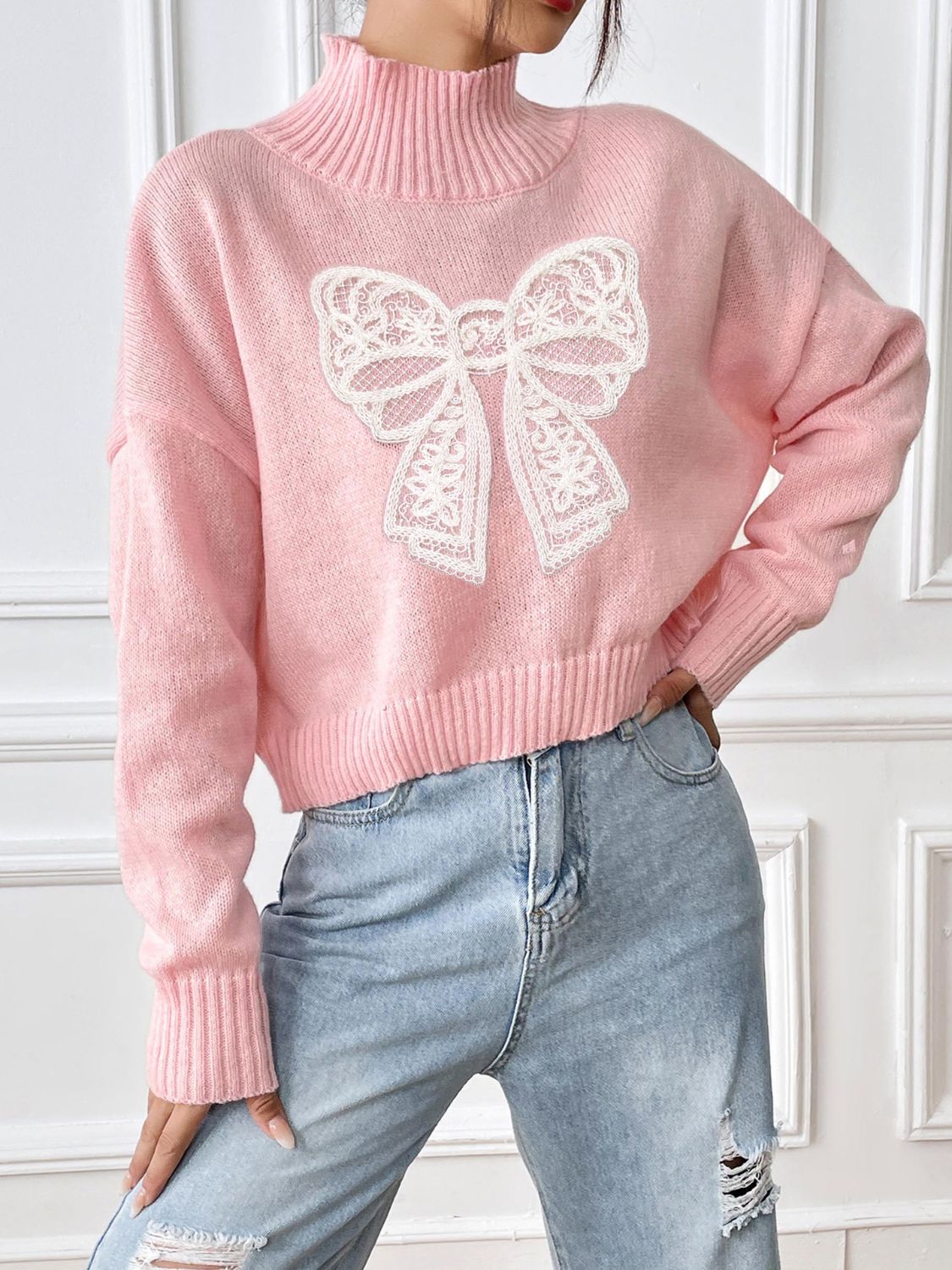 Pink turtleneck knit sweater with white lace bow appliqué and ribbed cuffs.