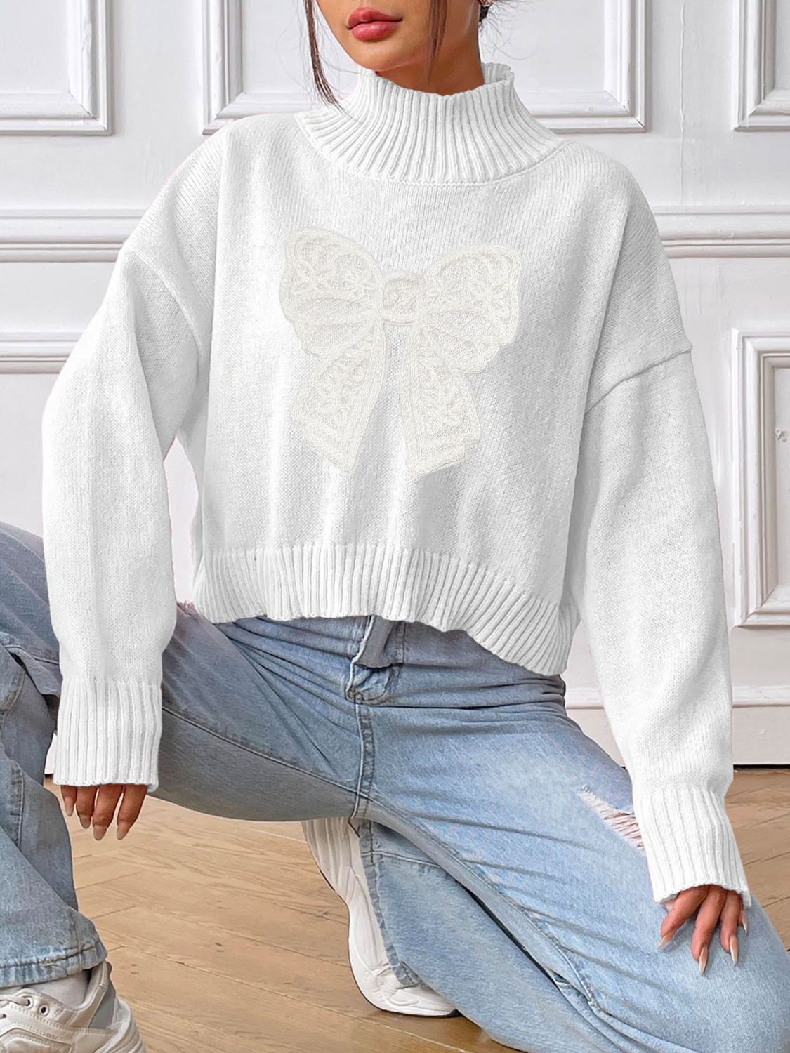 White turtleneck knit sweater with lace bow appliqué detail.